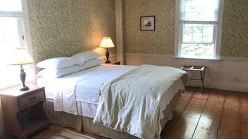 Une chambre à coucher avec un papier peint à motifs floraux, un lit recouvert de draps blancs, une table de chevet avec une lampe et une fenêtre avec des stores.