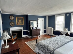 A bedroom with a fireplace, a bed with a floral bedspread, wooden furniture, and a view of the outdoors through a window. - Colonel Williams Inn (Brattleboro)