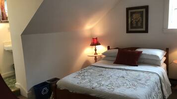 Une chambre avec un plafond en pente, un lit avec une literie blanche et un oreiller rouge, une table de chevet avec une lampe, et un tableau encadré au mur.