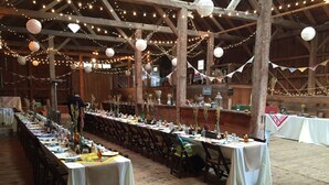 Una larga mesa de banquete dispuesta en un granero rústico, con luces de cuerda y farolillos de papel.