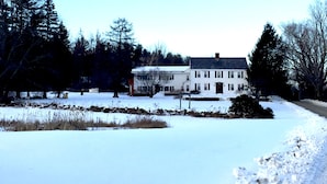 Una casa grande de color blanco con techo oscuro, rodeada de nieve en el suelo y árboles.