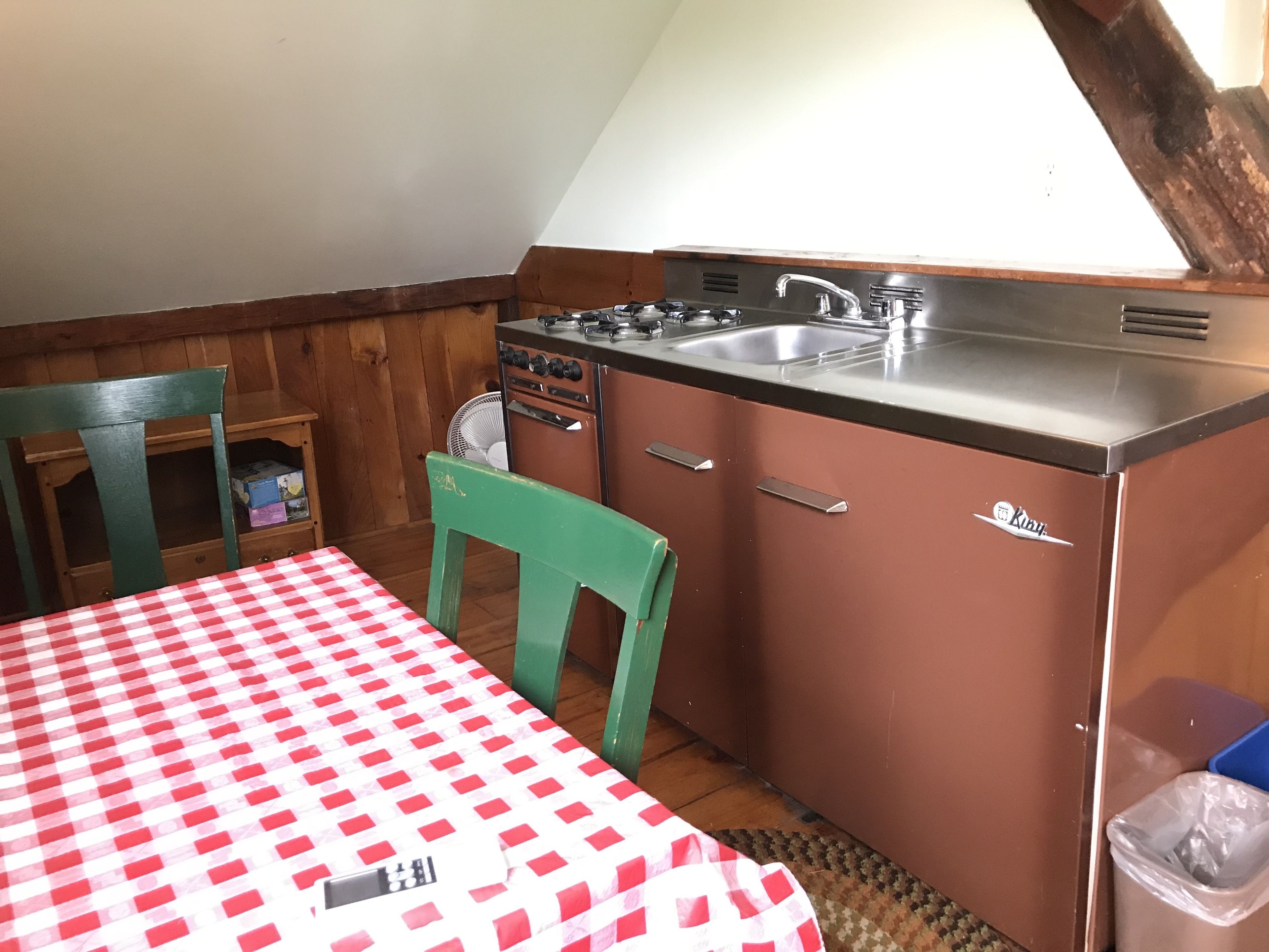 A compact kitchen with a red and white checkered tablecloth, a green chair, and a small fan.