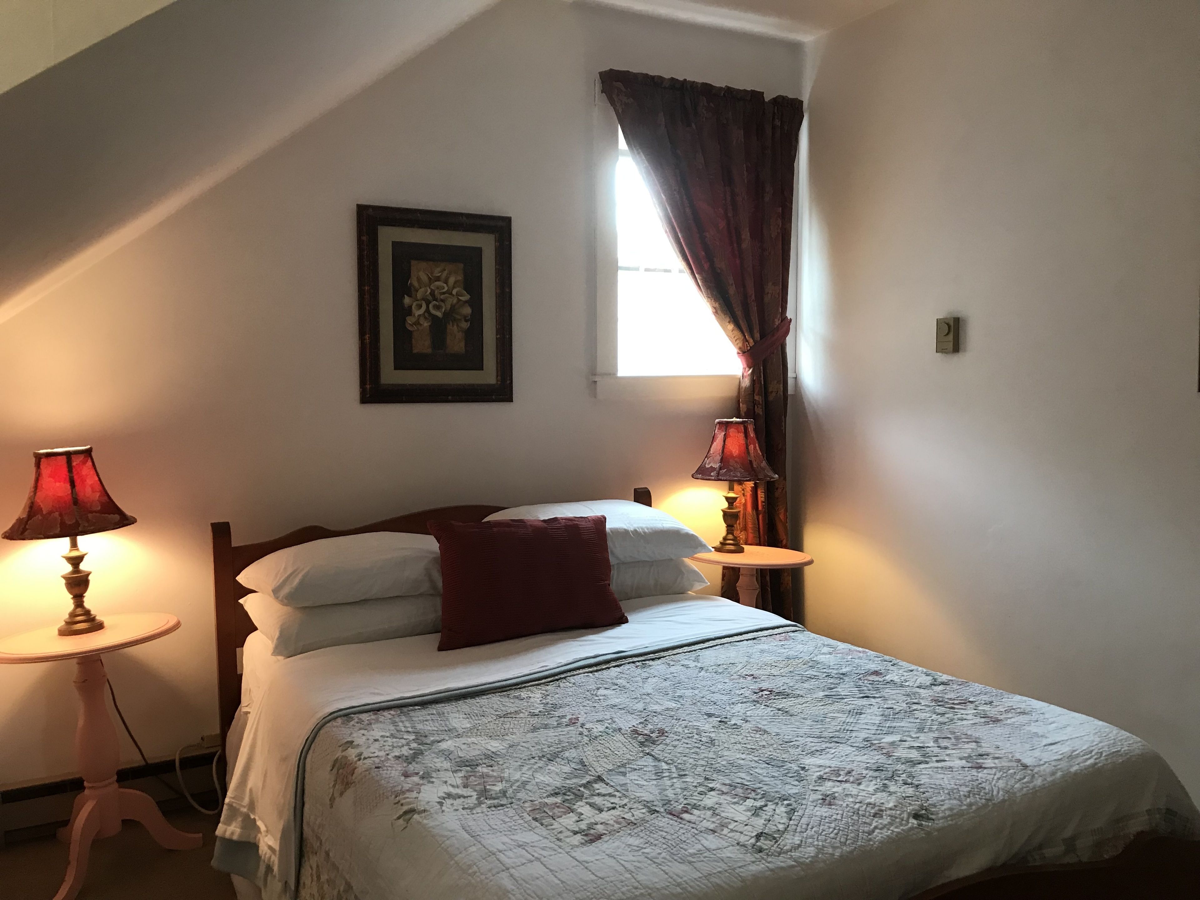 A bedroom with a bed, two bedside tables, a lamp, a window with curtains, and a framed picture on the wall.