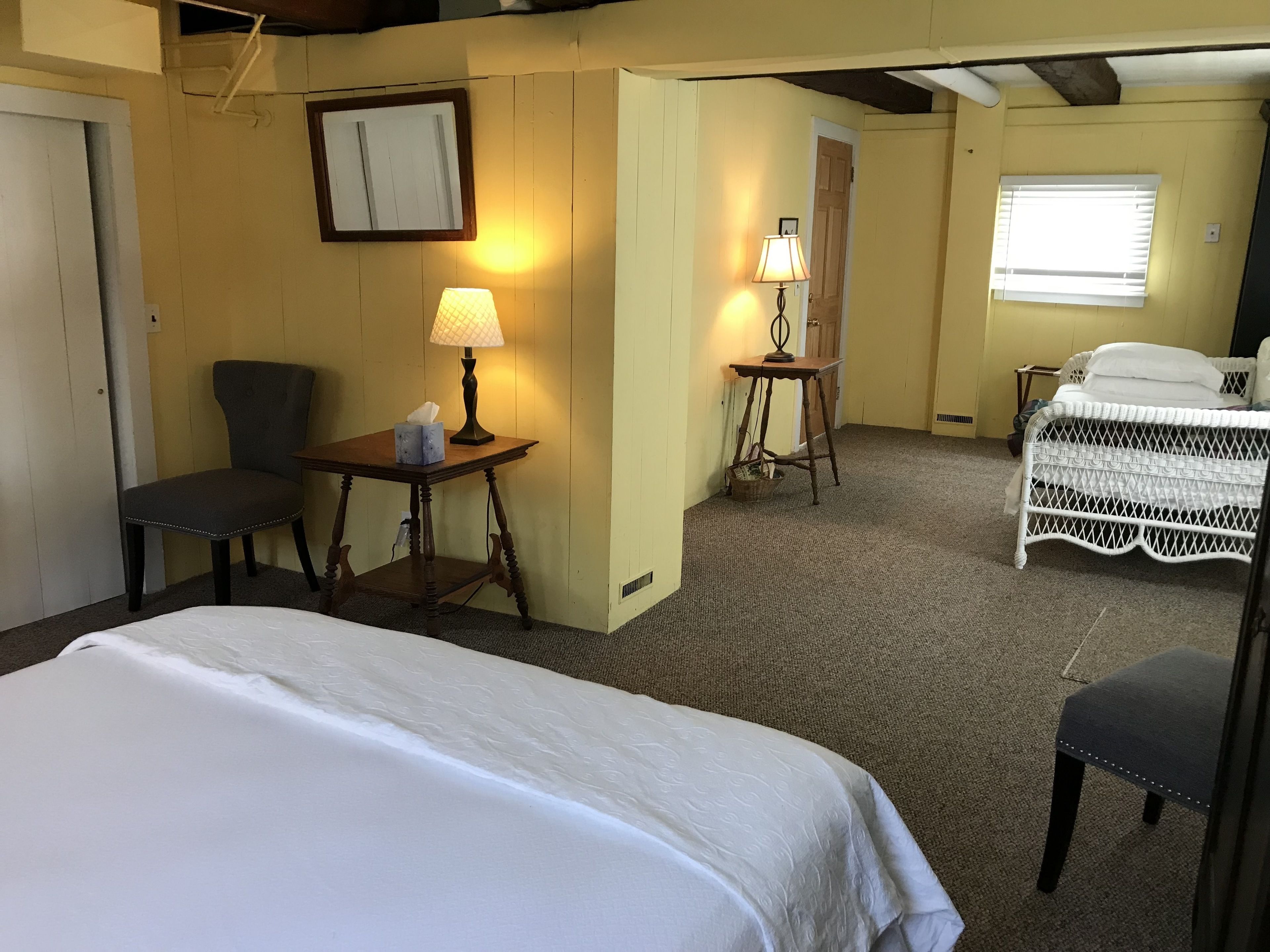 A hotel room with a bed, two chairs, a table with a lamp, a small table with a basket, and a window with blinds.