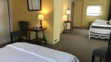 A hotel room with a bed, two chairs, a table with a lamp, a small table with a basket, and a window with blinds.