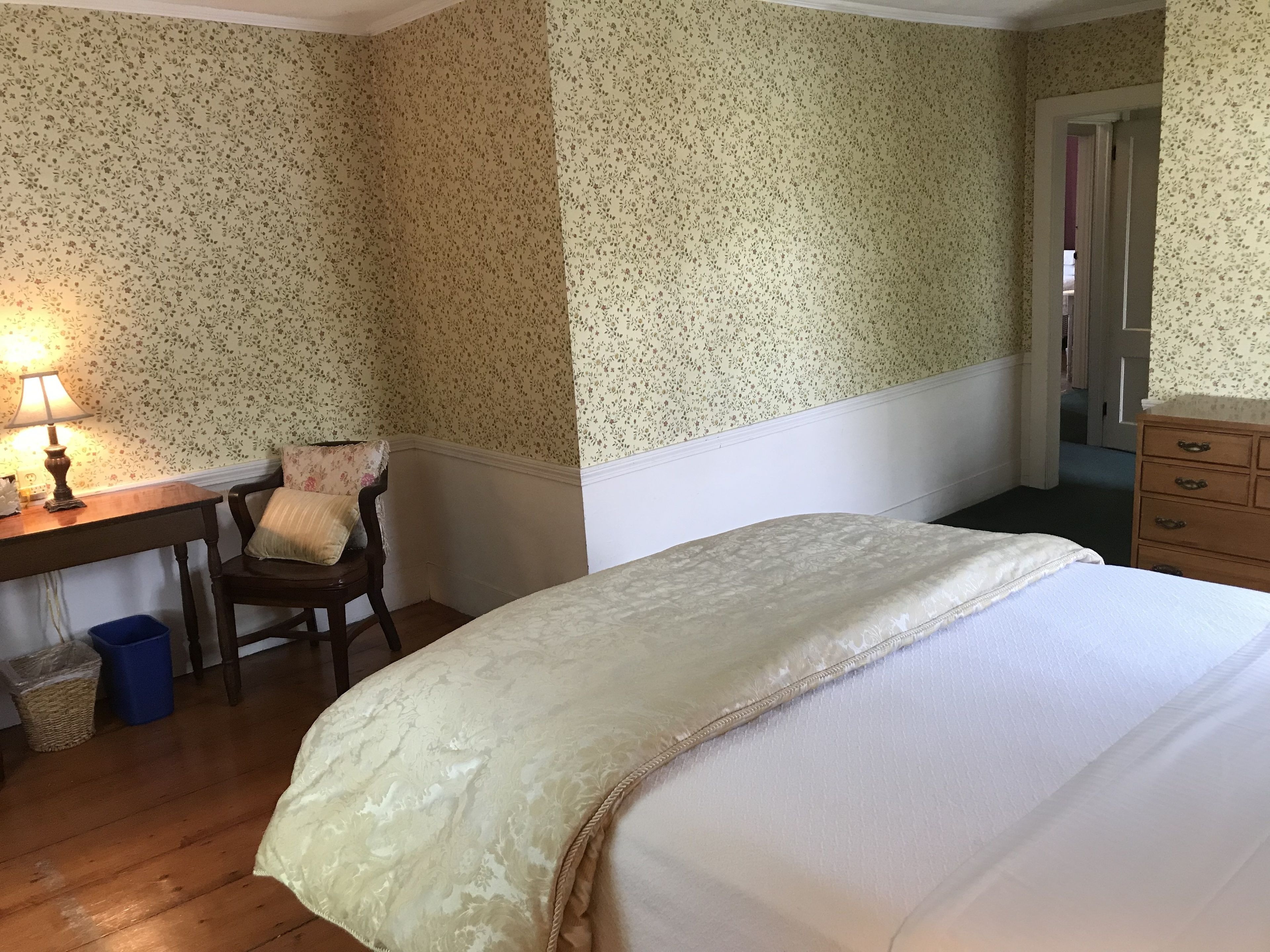 Une chambre avec un papier peint à motifs floraux, un bureau en bois avec une lampe, une chaise, un lit recouvert d’une couverture blanche et une commode.
