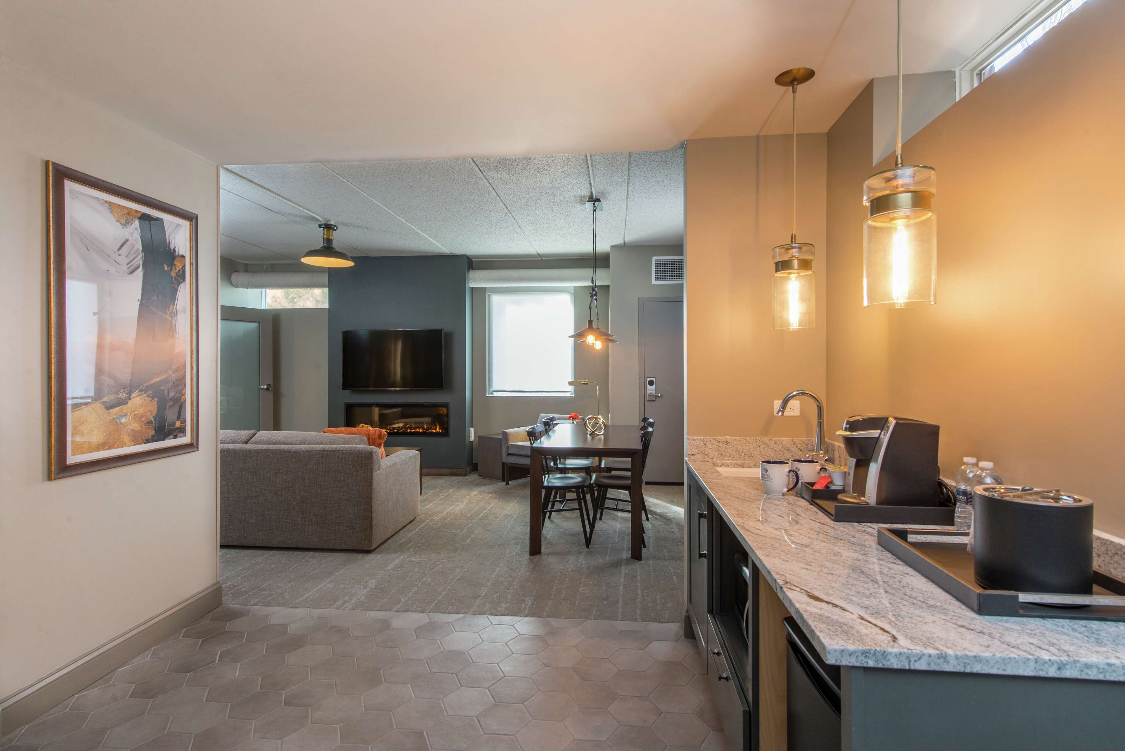 A modern kitchen with a marble countertop, a built-in oven, and a sink. There is a dining area with a table and chairs, and a living room with a sofa and a TV.