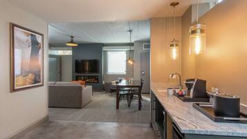 A modern kitchen with a marble countertop, a built-in oven, and a sink. There is a dining area with a table and chairs, and a living room with a sofa and a TV.