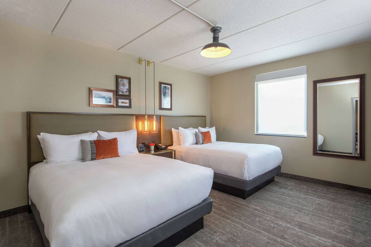 A hotel room with two beds, a large window, a mirror, and framed pictures on the wall.