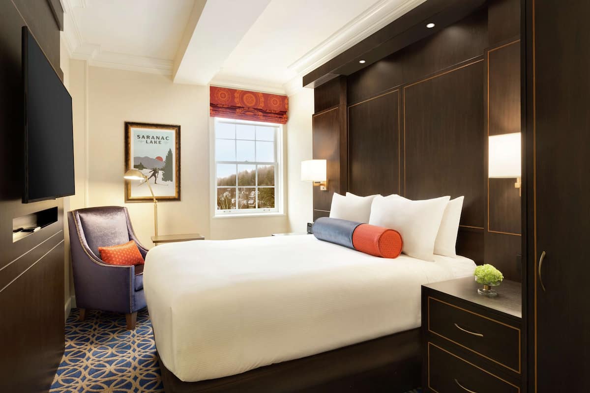 A hotel room with a large bed, a chair, a TV, a window with a view, and a framed picture on the wall.