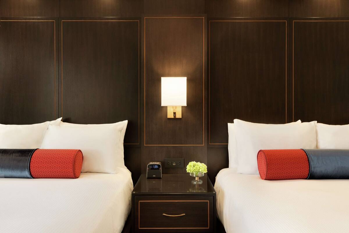 A hotel room with two beds, a nightstand with a lamp, and a small table with a clock and a vase of flowers.