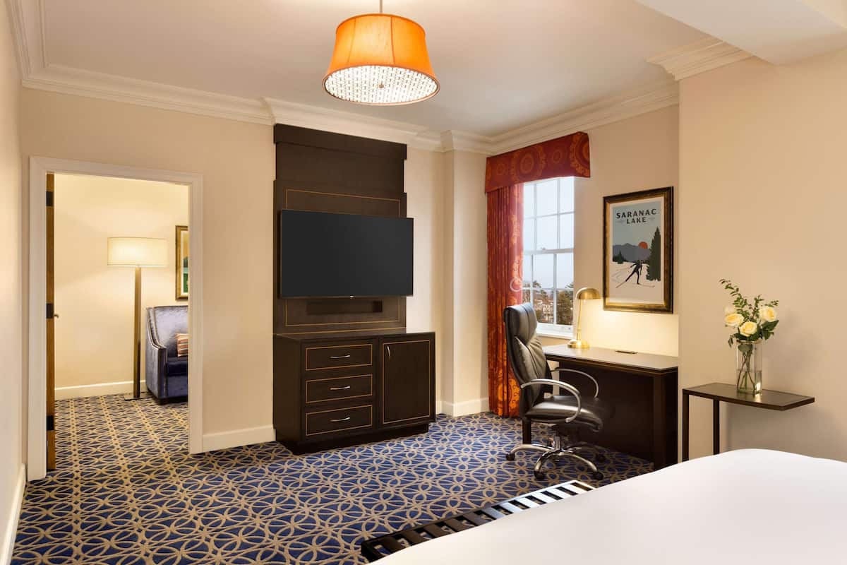 A hotel room with a patterned carpet, a bed, a desk with a chair, a television, a window with curtains, and a framed picture on the wall.
