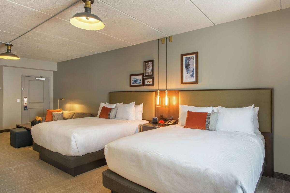 A hotel room with two beds, a bedside table, a lamp, and a door.