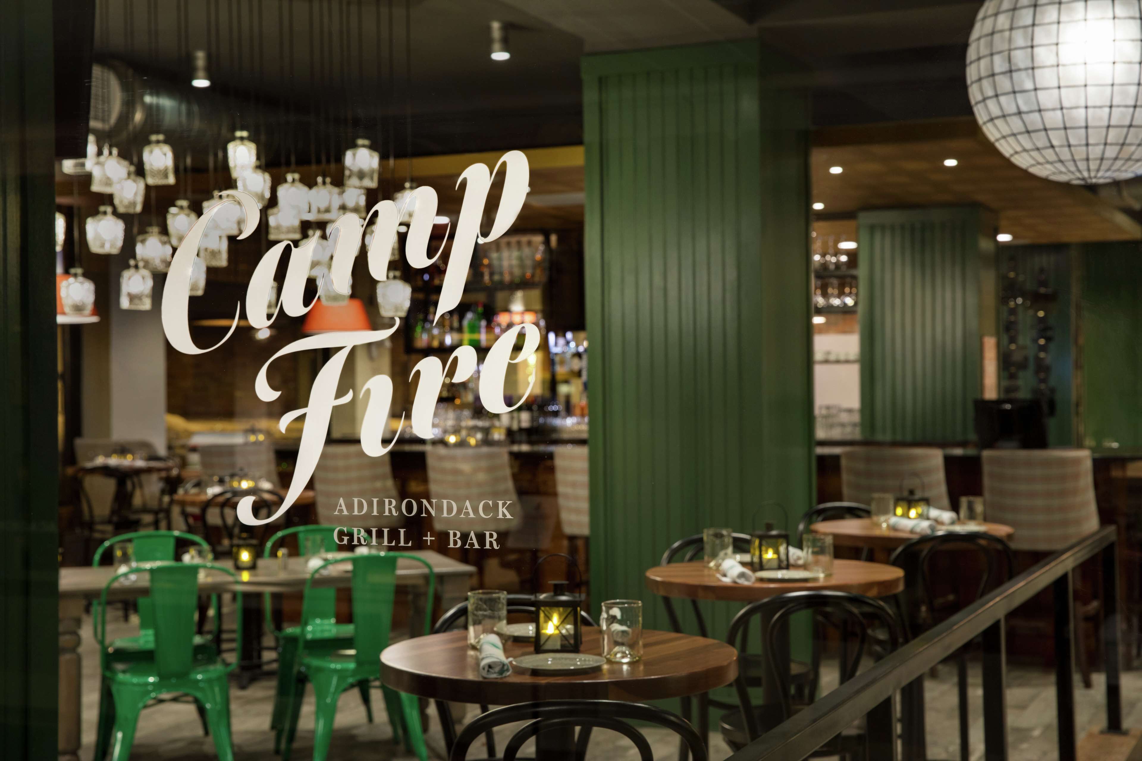An interior view of Adirondack Grill + Bar with round tables, green chairs, and hanging lanterns.