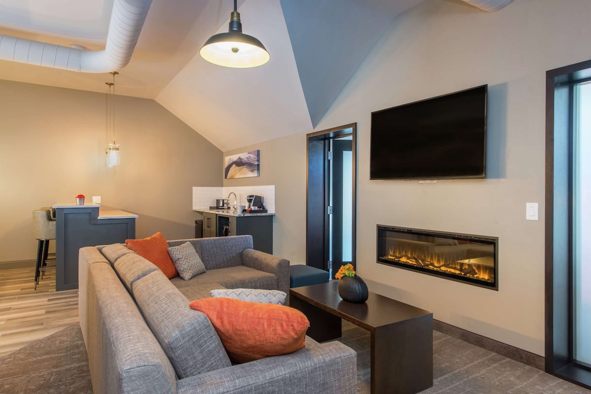 A modern living room with a sectional sofa, a fireplace, a TV mounted on the wall, and a kitchen area in the background.
