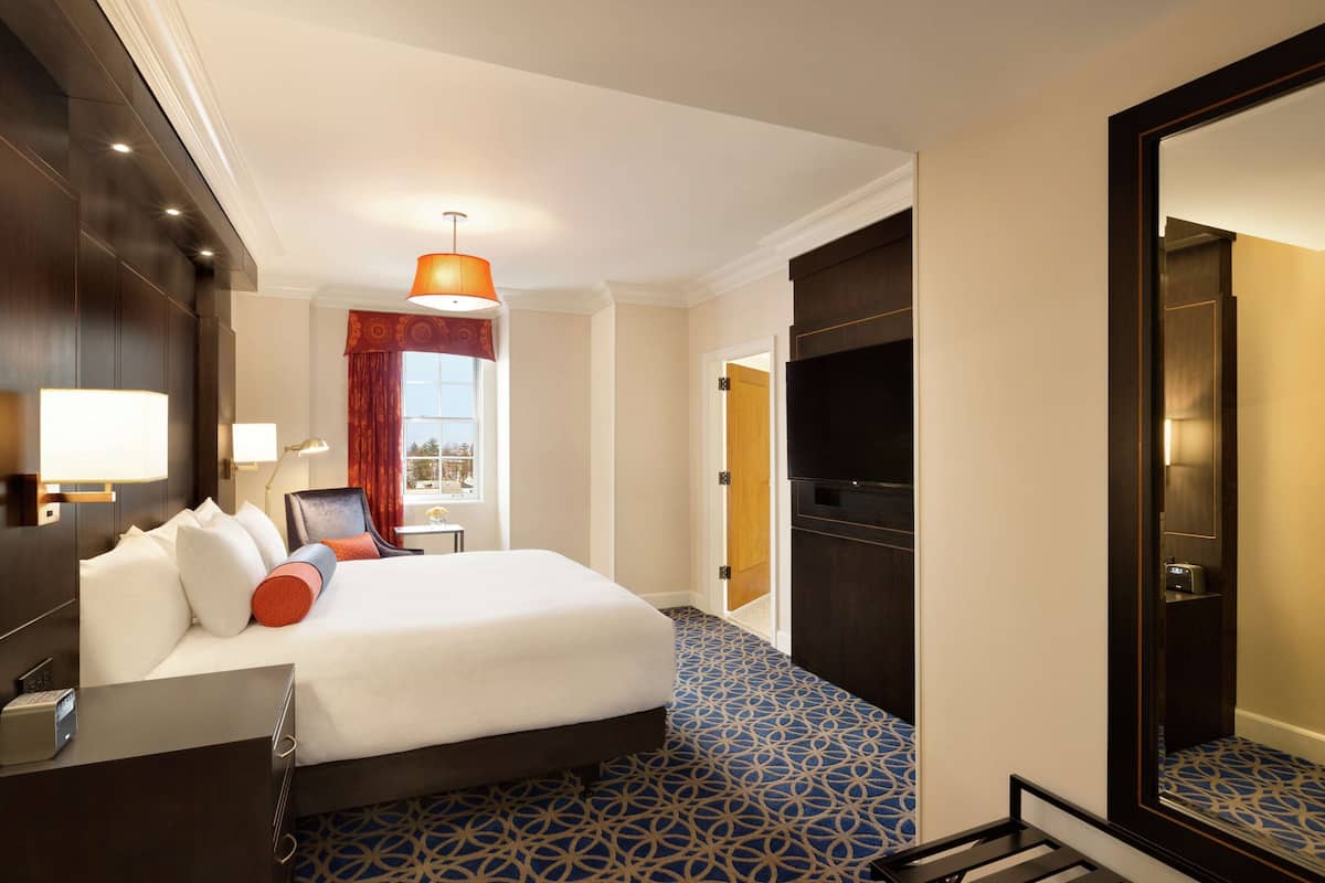 A hotel room with a large bed, a desk with a lamp, a TV, and a patterned carpet.