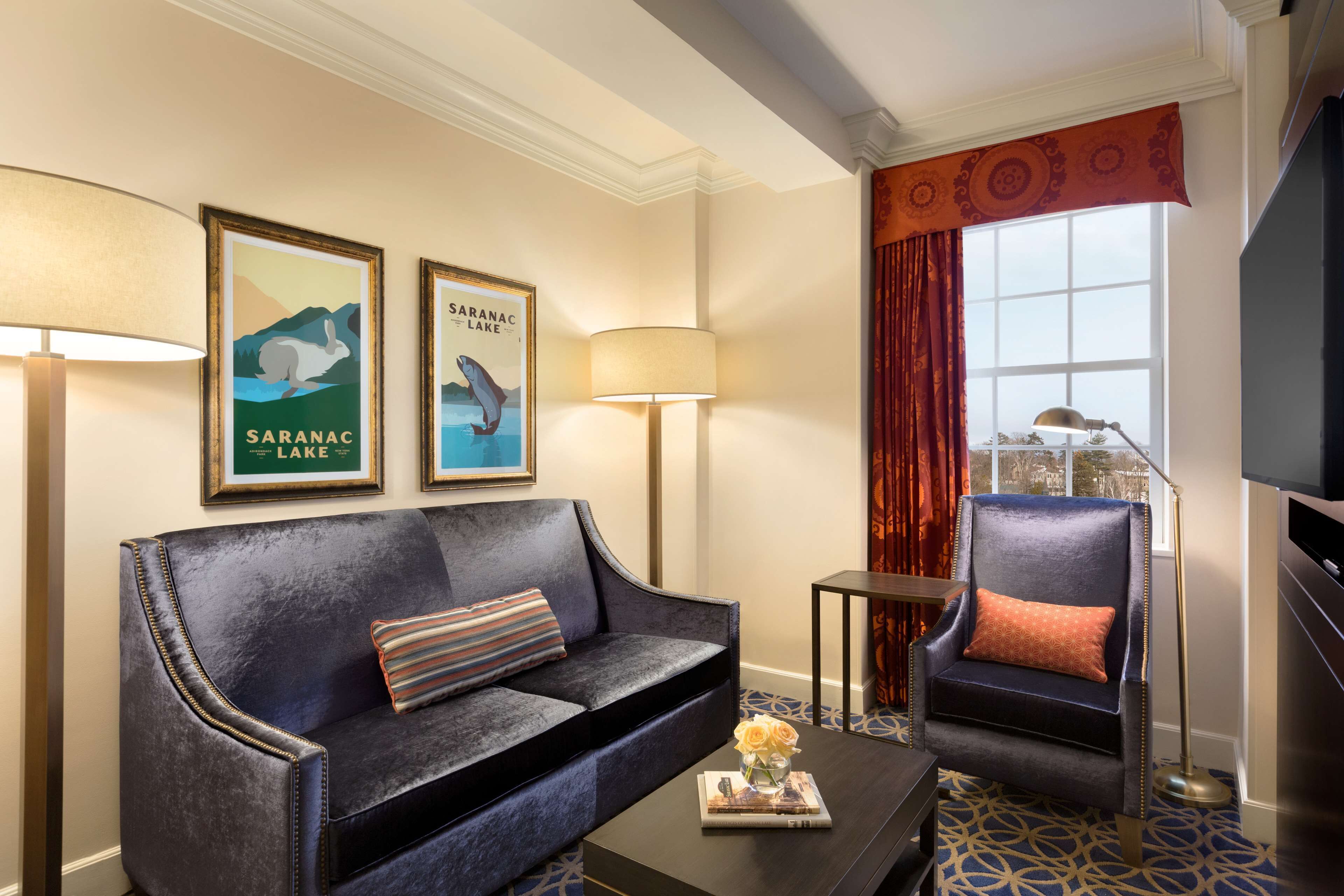 A modern hotel room with a dark blue sofa, a matching armchair, a coffee table with a vase of flowers, and two framed posters on the wall.