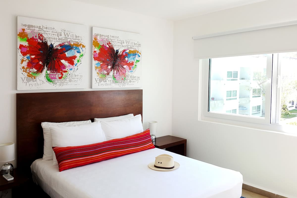A hotel room with a large bed, two wall art pieces, a window with a view of buildings, and a hat on the bed.
