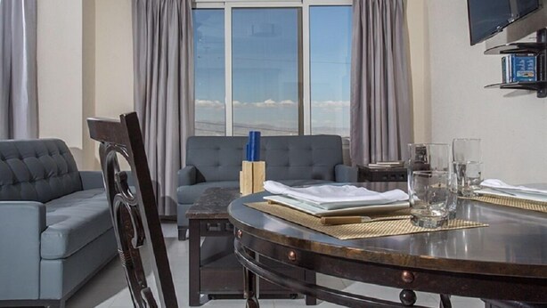 A modern living room with a grey sofa, a dining table with a glass top, and a view of the mountains through large windows. - Residences Talou (Petionville)