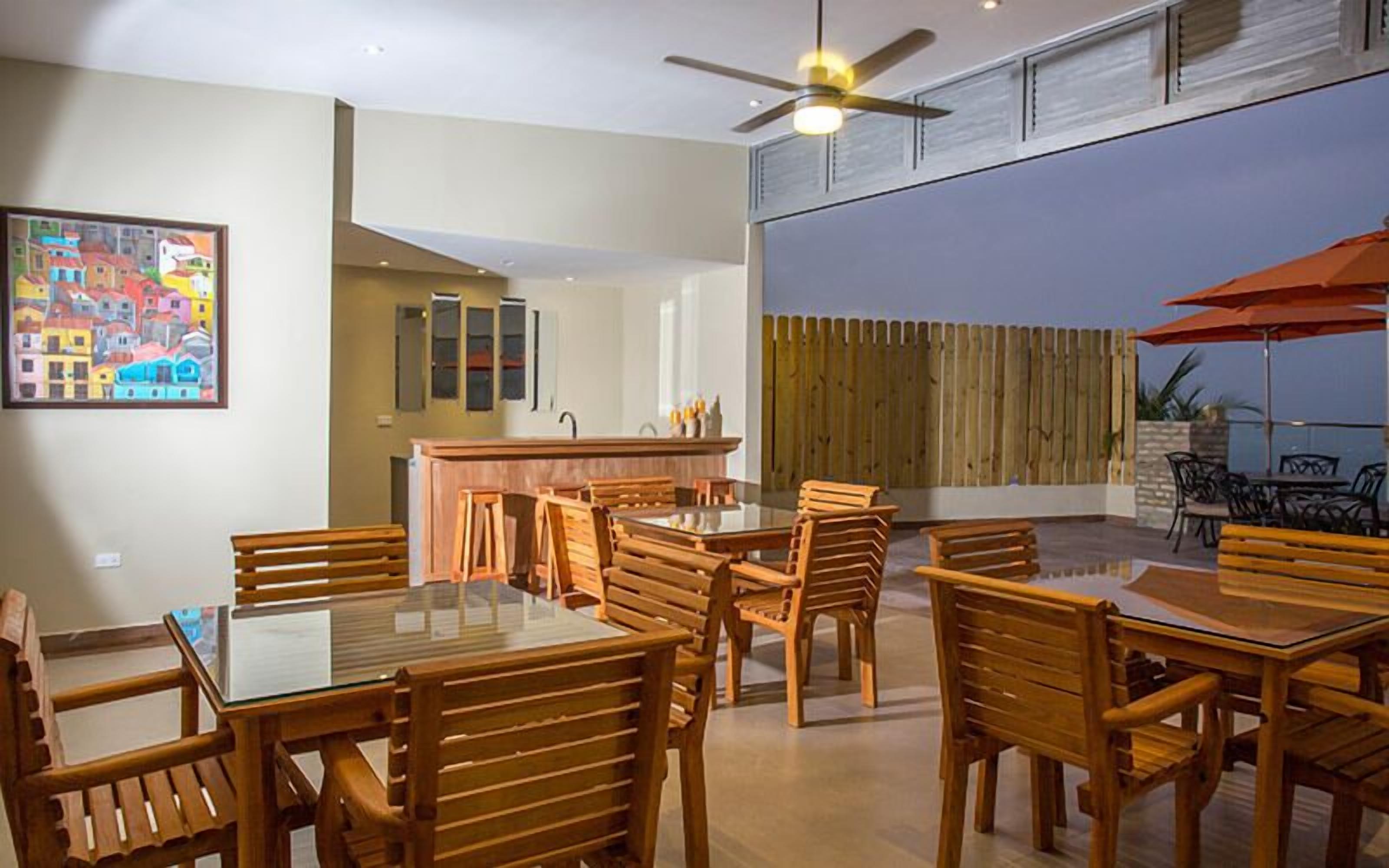 A dining area with wooden tables and chairs, a kitchen area with cabinets, and a large window with a view of a wooden fence.