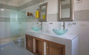 A bathroom with two glass sinks, wooden cabinets, a toilet, and a glass shower enclosure. - Residences Talou (Petionville)