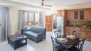 A spacious living area with a sofa, dining table, and chairs, a kitchen with wooden cabinets, and a view through large windows. - Residences Talou (Petionville)