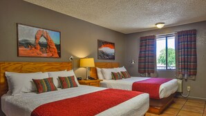 A hotel room with two beds, a wooden headboard, a nightstand with a lamp, a window with curtains, and two wall-mounted paintings.