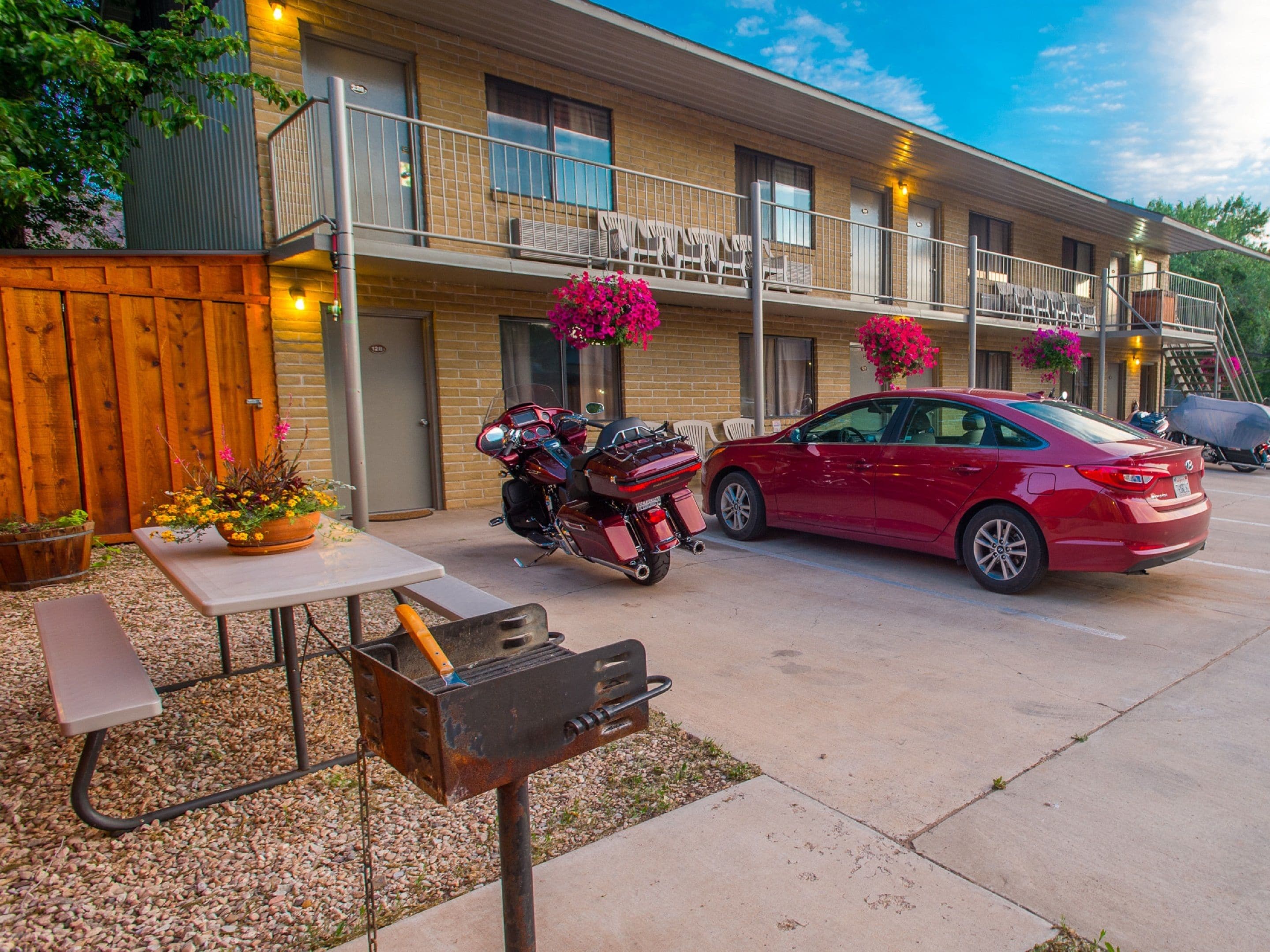 Ein zweistöckiges Motel mit überdachtem Parkplatz, einem Grill und einem roten Auto davor.