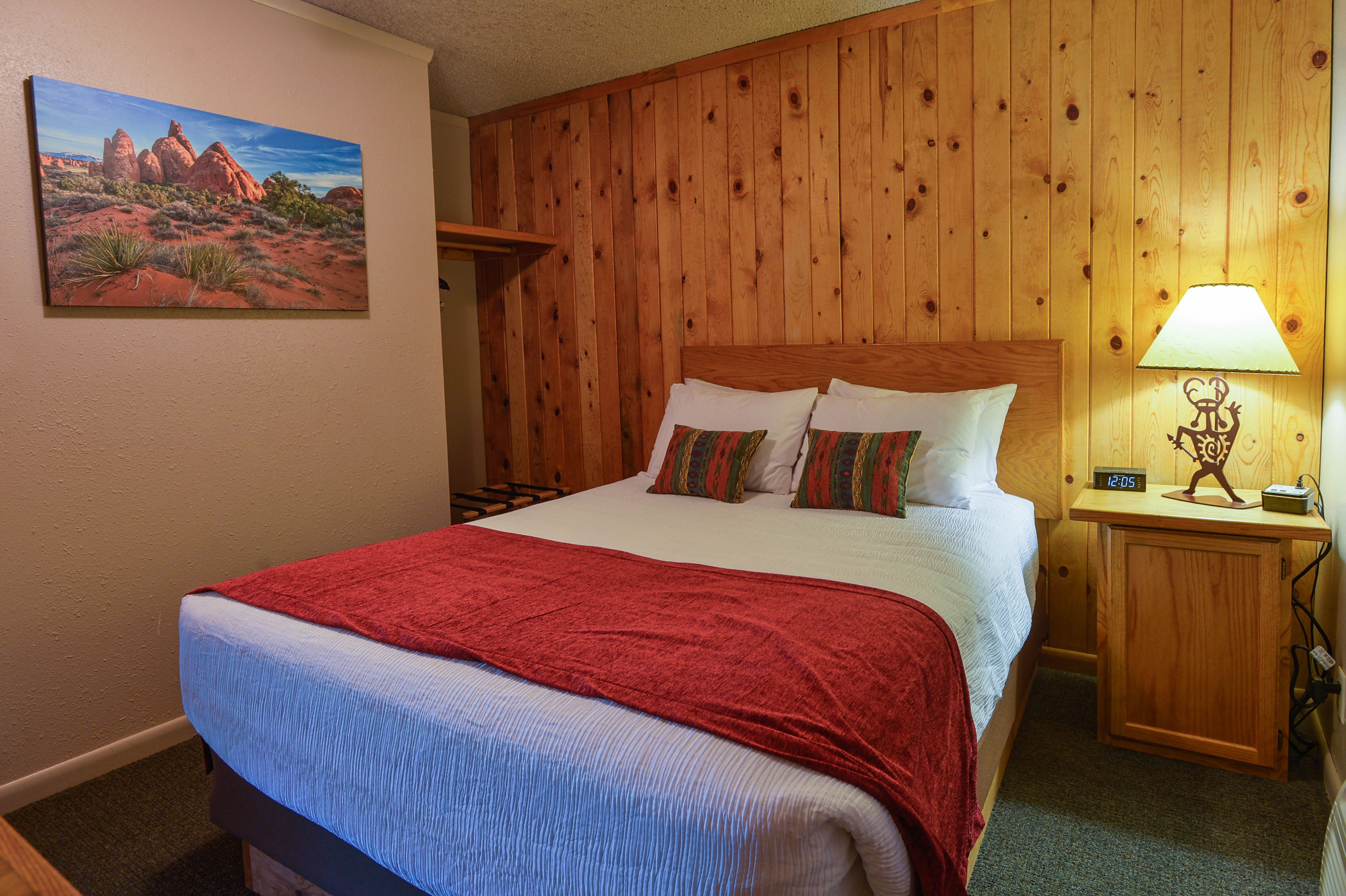 A wooden-paneled room with a bed, a nightstand with a lamp, and a painting of a desert landscape.