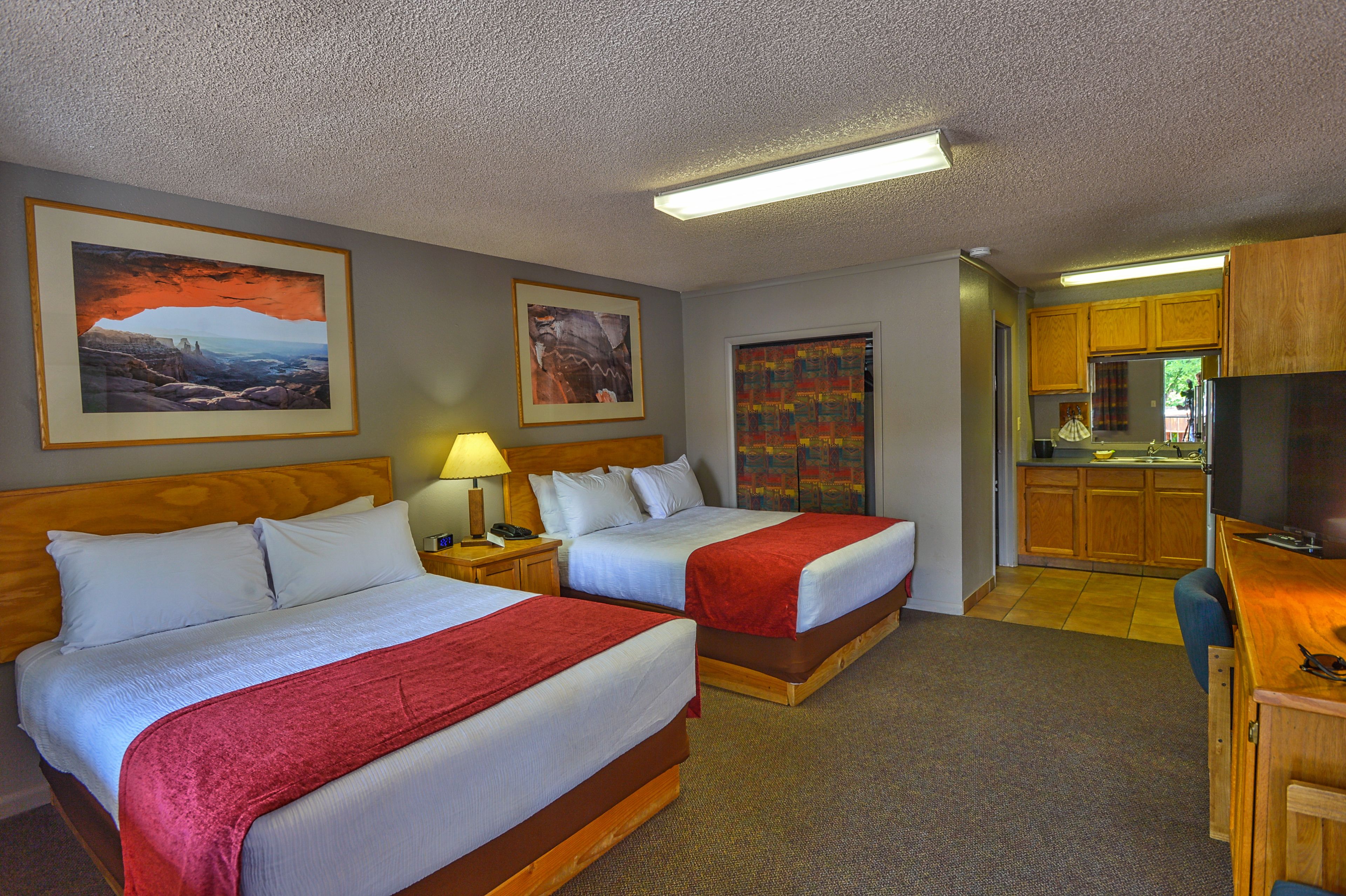 A hotel room with two beds, a desk, a kitchenette, and artwork on the walls.