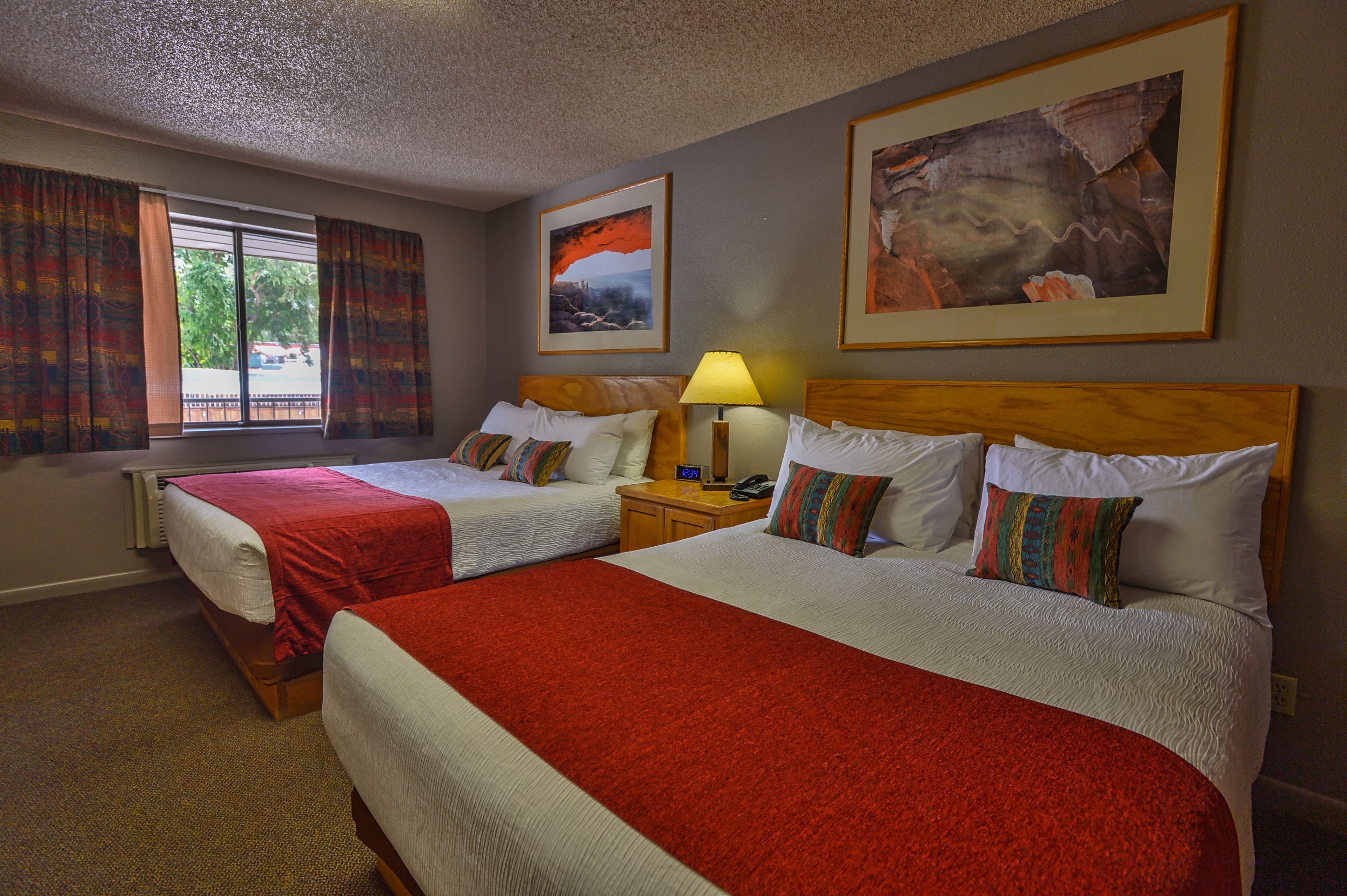 A hotel room with two beds, a window with curtains, a lamp, and artwork on the walls.