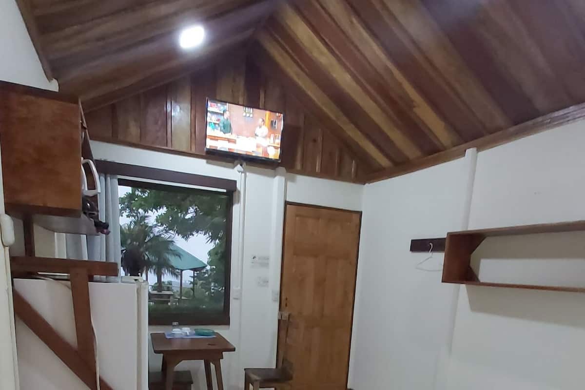 A compact room with a bed, a small table, a chair, a TV mounted on the wall, and a door.