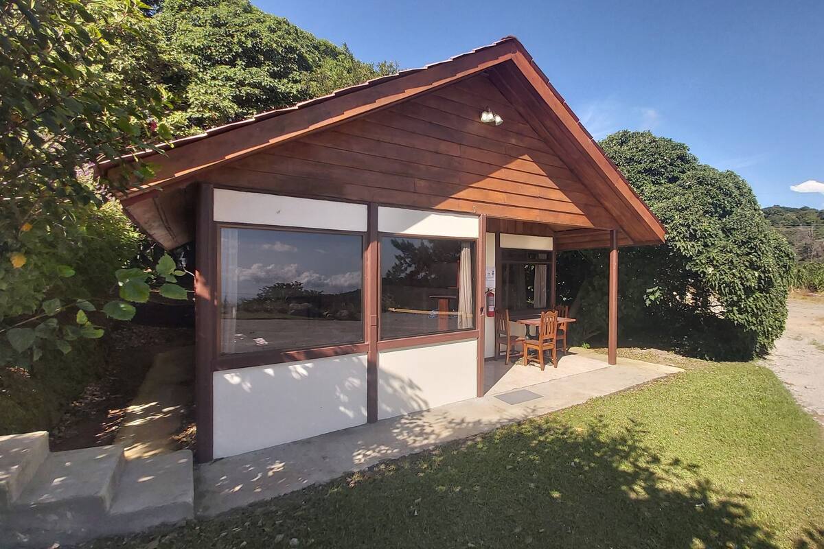A wooden cabin with large windows, a patio with chairs, and a view of the surrounding greenery.