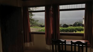 Una habitación con una mesa de madera y dos ventanas con cortinas, que da a un jardín con rocas y follaje.