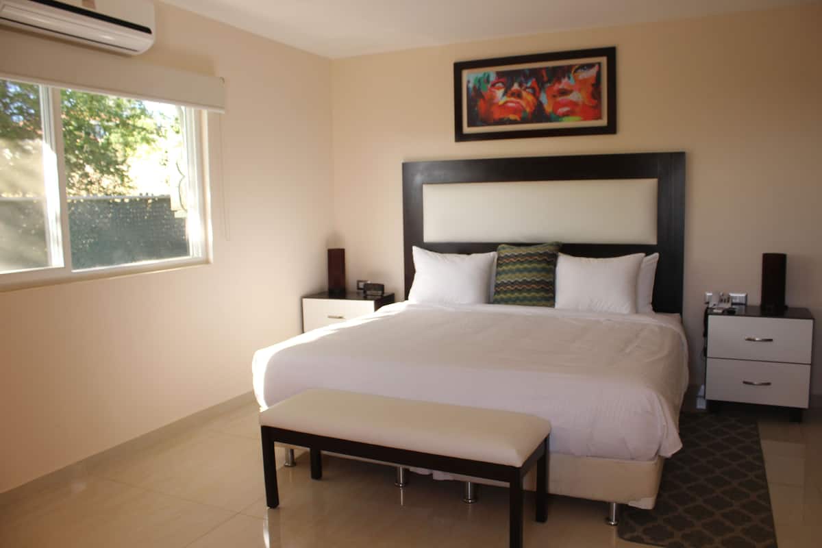 A hotel room with a large bed, a bench, a nightstand, a bedside table, and a painting on the wall.