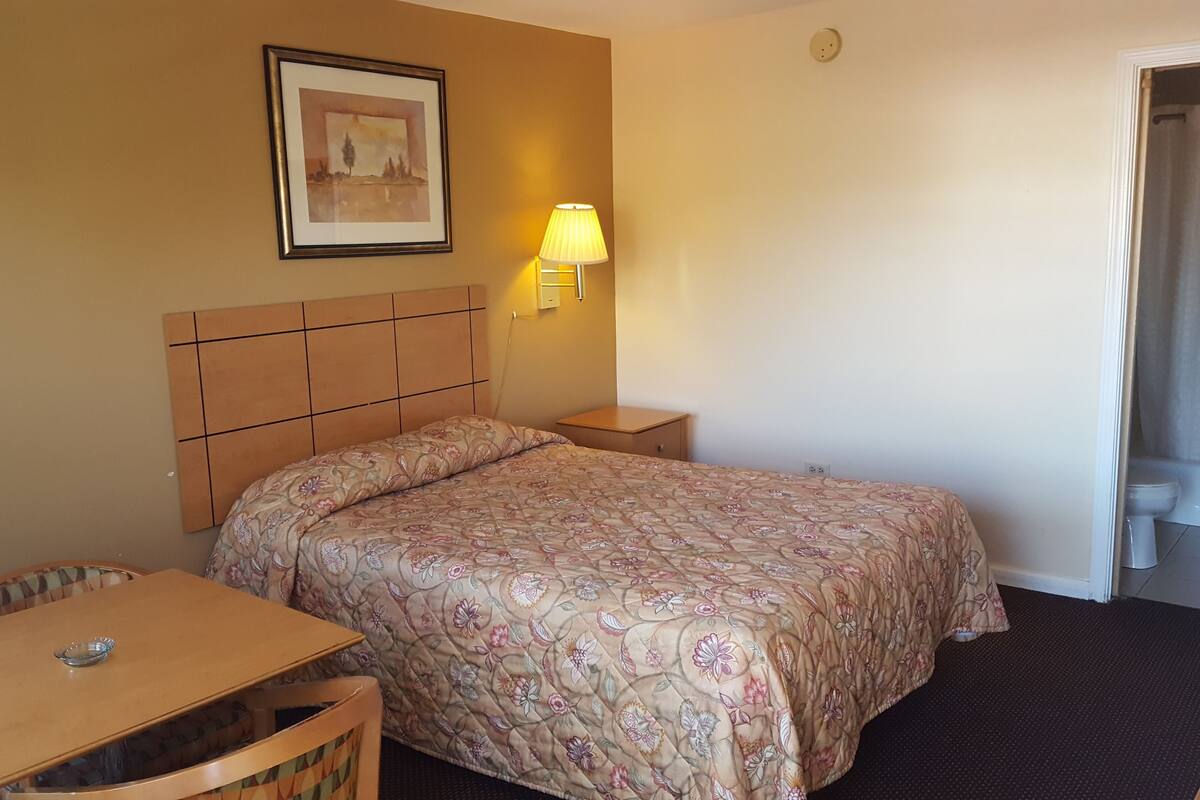 Une chambre d’hôtel avec un lit, un bureau, une chaise, une table de chevet, une lampe et un tableau encadré au mur.