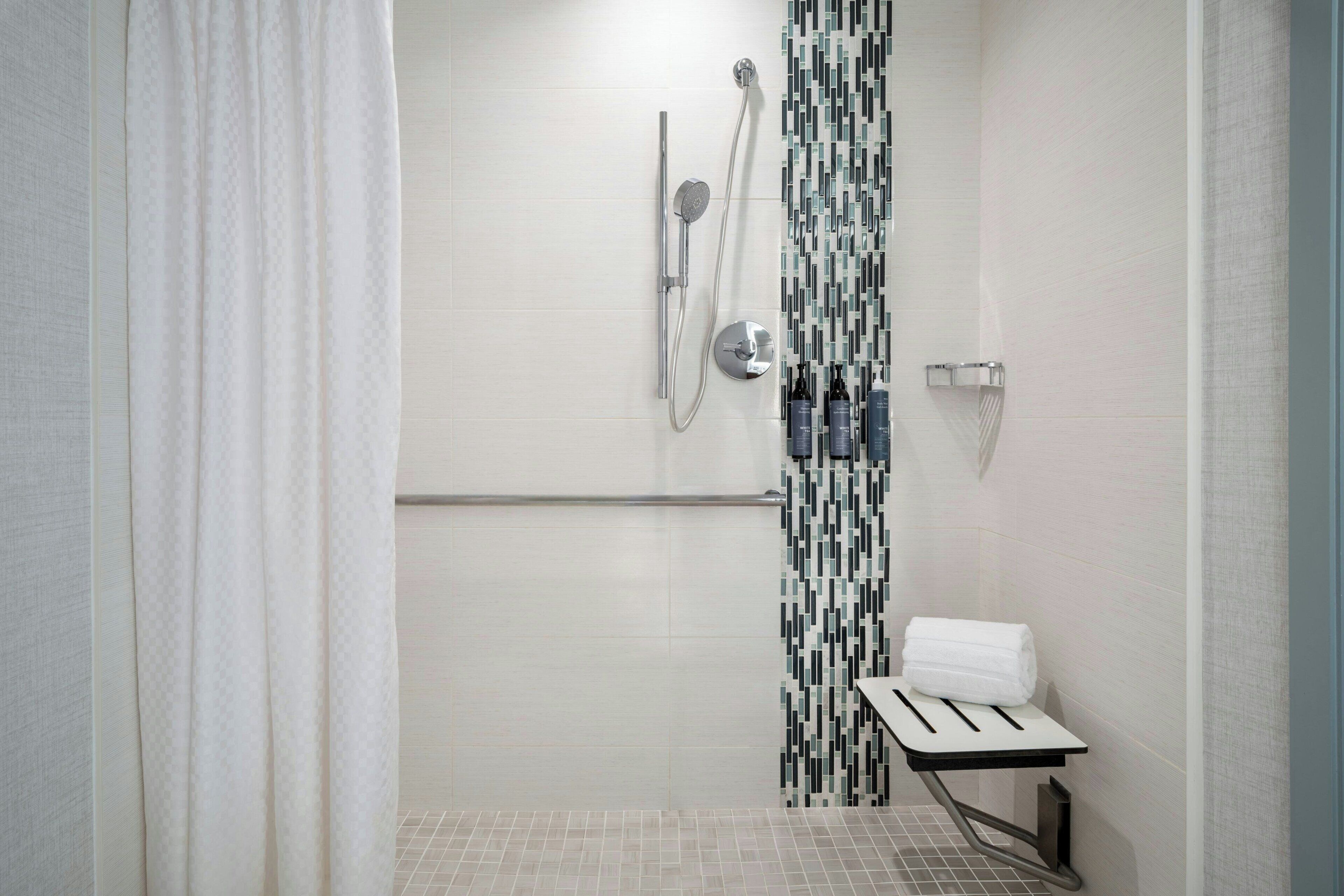A modern shower with a handheld showerhead and a wall-mounted faucet. There is a towel rack with a folded towel and a bottle of shampoo.