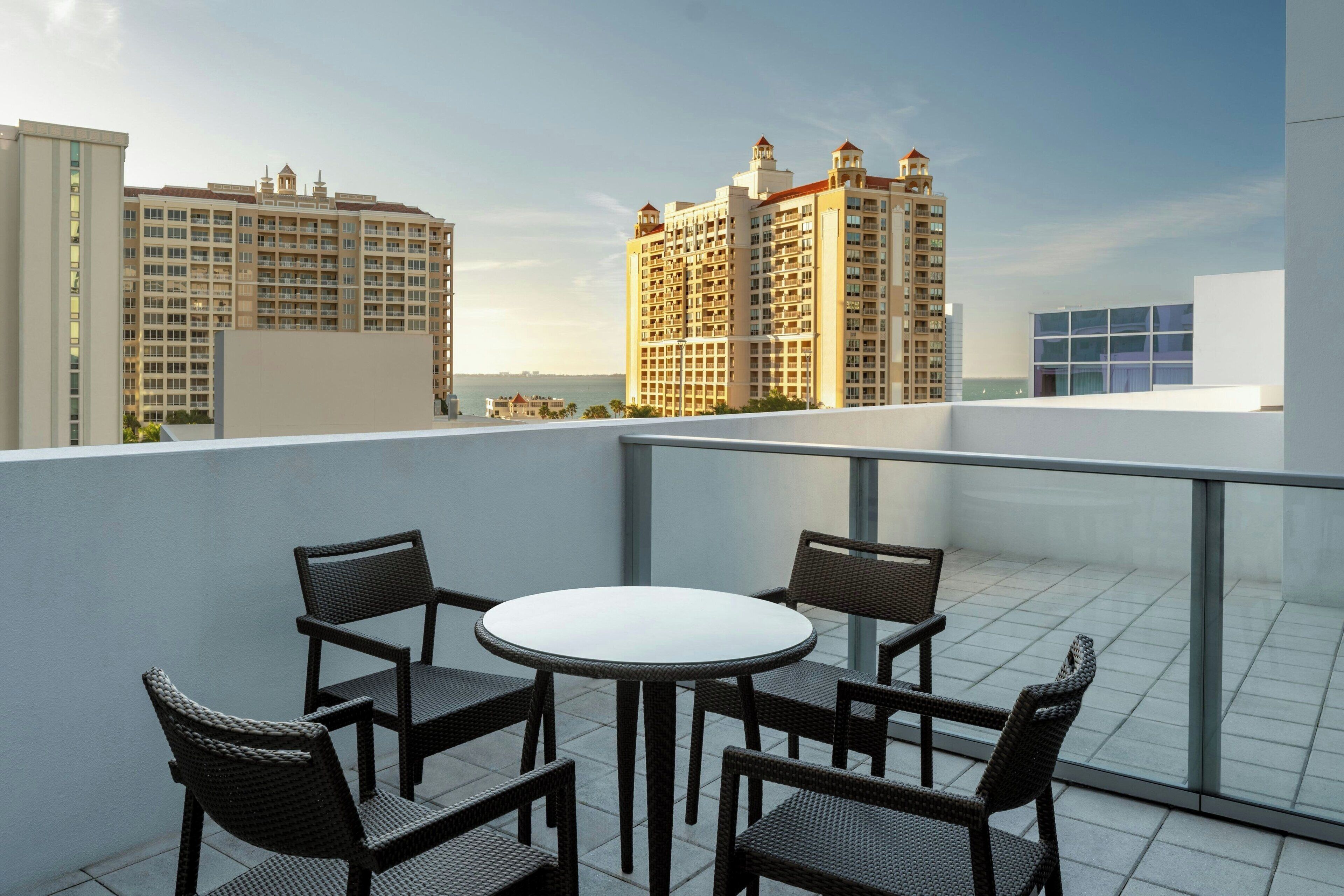 A rooftop terrace with a round table and four wicker chairs, overlooking a cityscape with high-rise buildings.