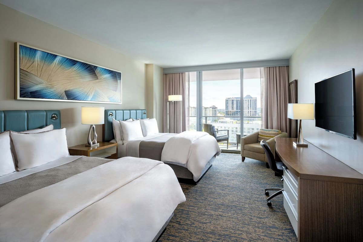 Ein Hotelzimmer mit zwei Betten, einem Schreibtisch, einem Fernseher und einem großen Fenster mit Blick auf die Stadt.