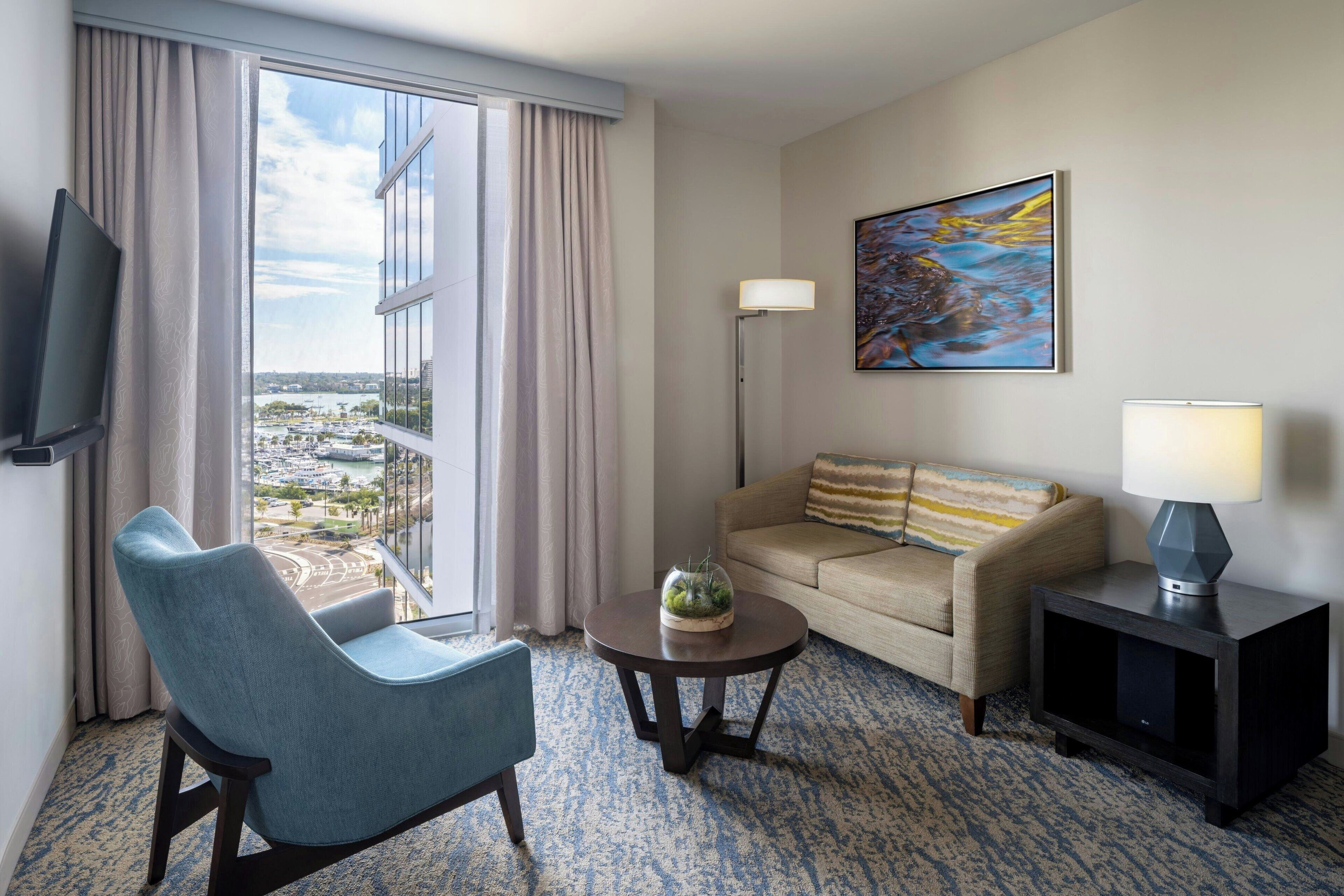 A hotel room with a view of a cityscape, a blue armchair, a beige sofa, a small table with a lamp, and a painting on the wall.