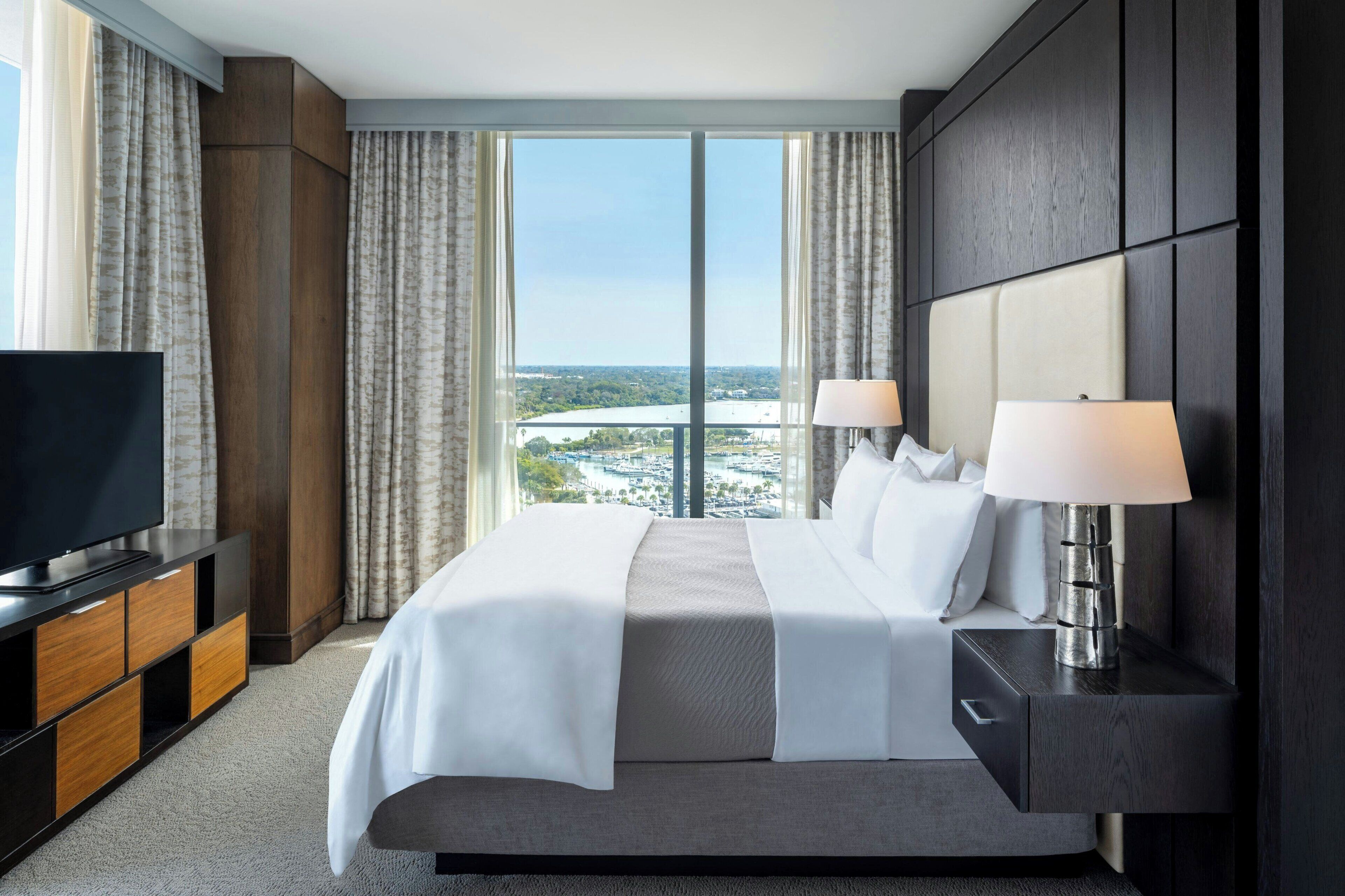 A hotel room with a large bed, two bedside lamps, a TV, and a view of a cityscape.