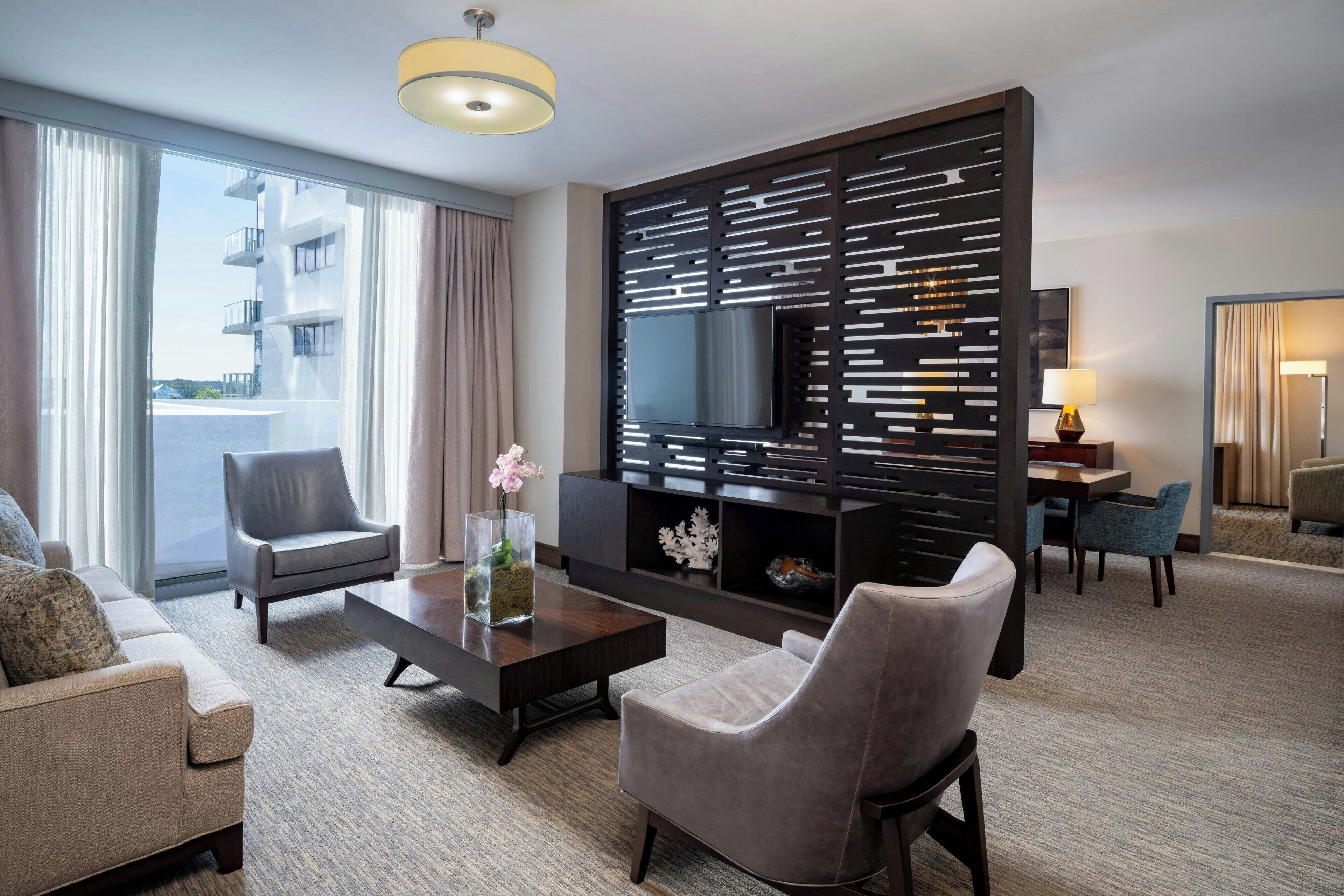 A modern hotel room with a large window, a flat-screen TV, a sofa, a coffee table, and a decorative wall panel.