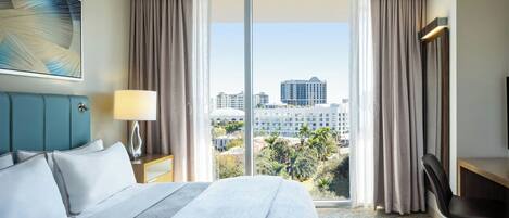 A hotel room with a large bed, a desk, a chair, a lamp, and a view of the city through large windows.