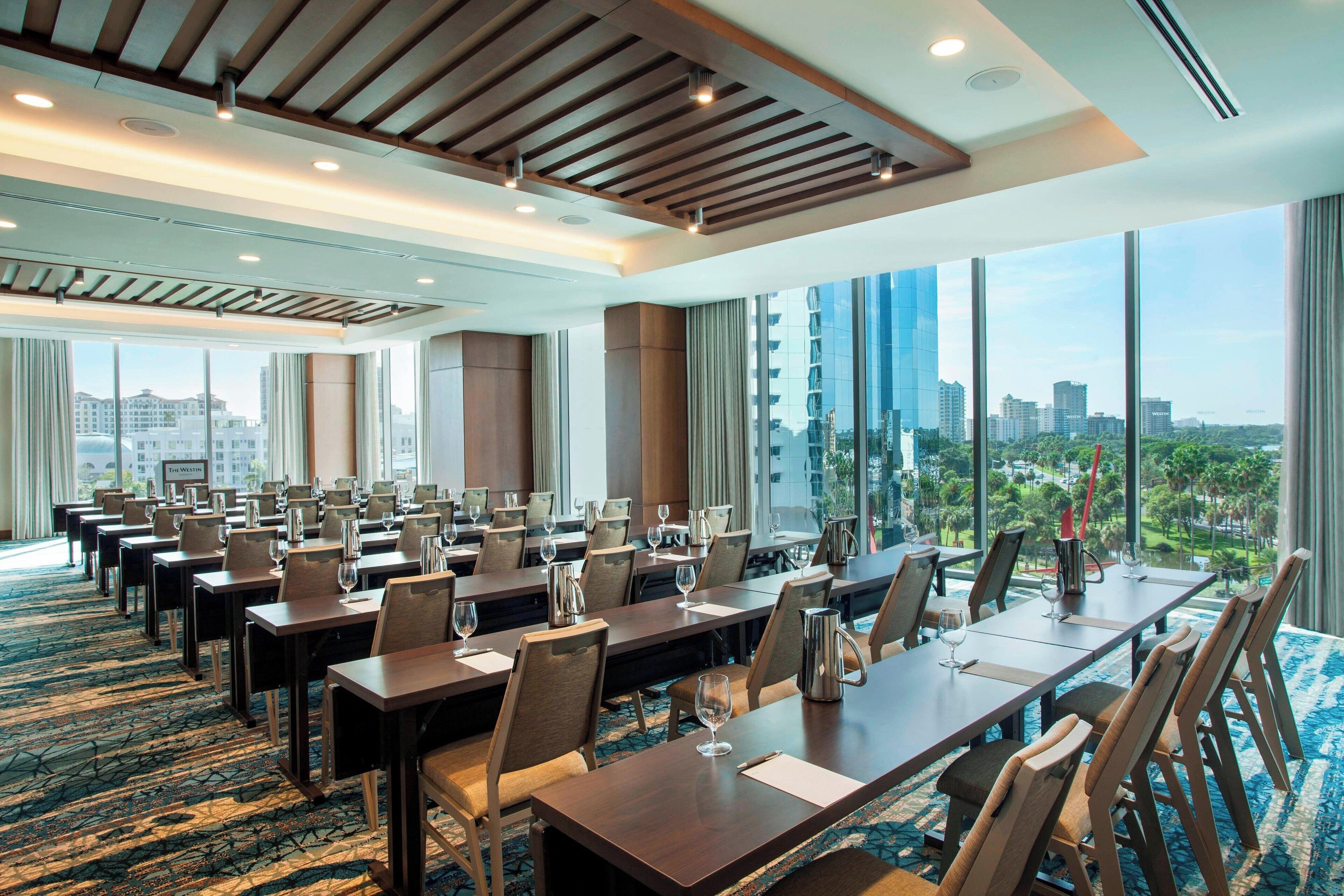 A conference room with multiple tables and chairs, large windows with a city view, and a patterned carpet.