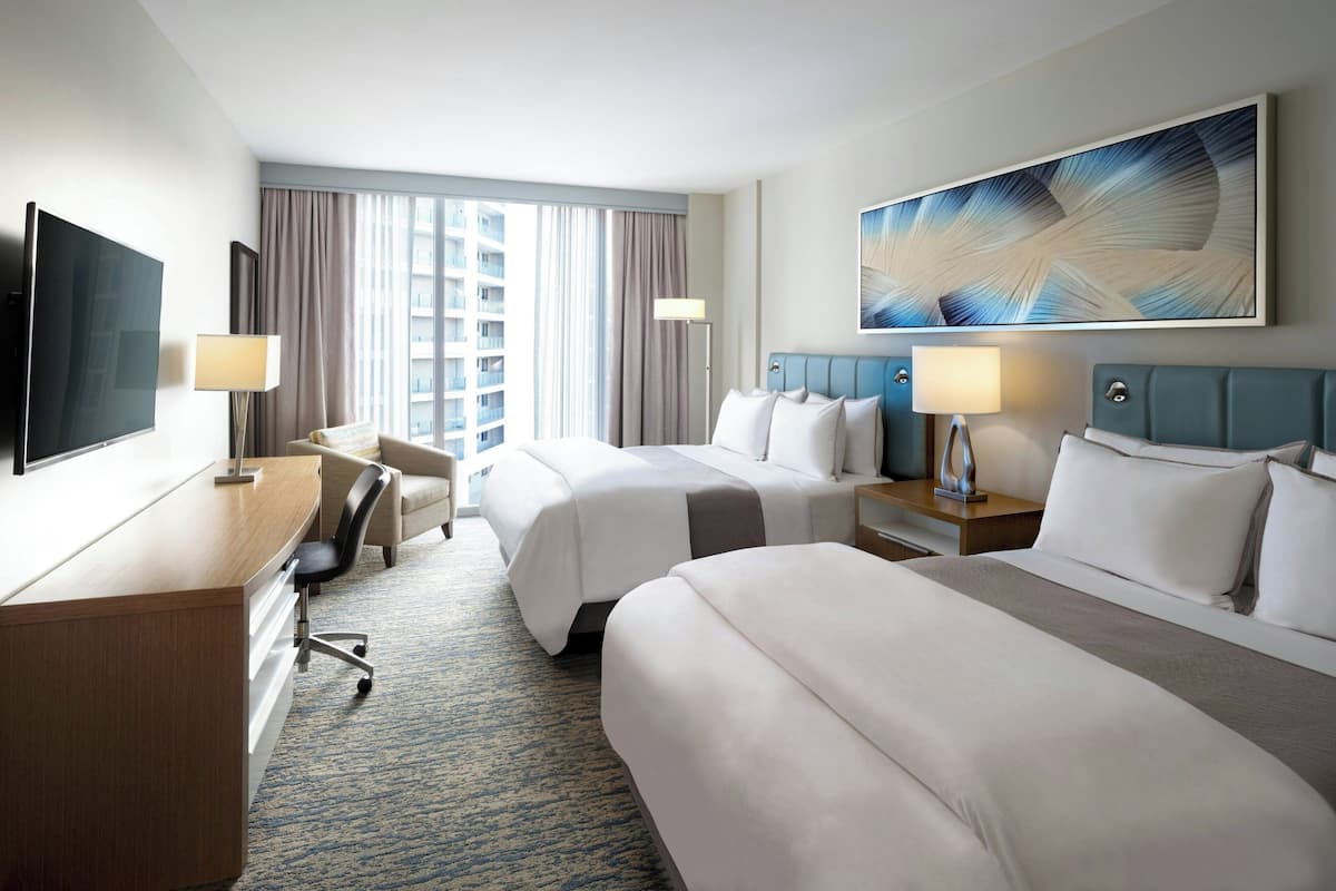 Ein Hotelzimmer mit zwei Betten, einem Schreibtisch, einem Stuhl, einem Fernseher und einem großen Fenster mit Vorhängen.