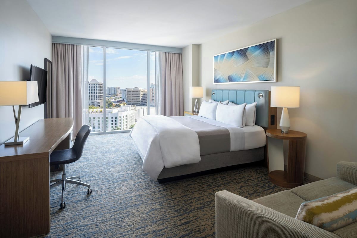 Ein Hotelzimmer mit einem großen Bett, einem Schreibtisch, einem Sessel und Blick auf die Stadt.