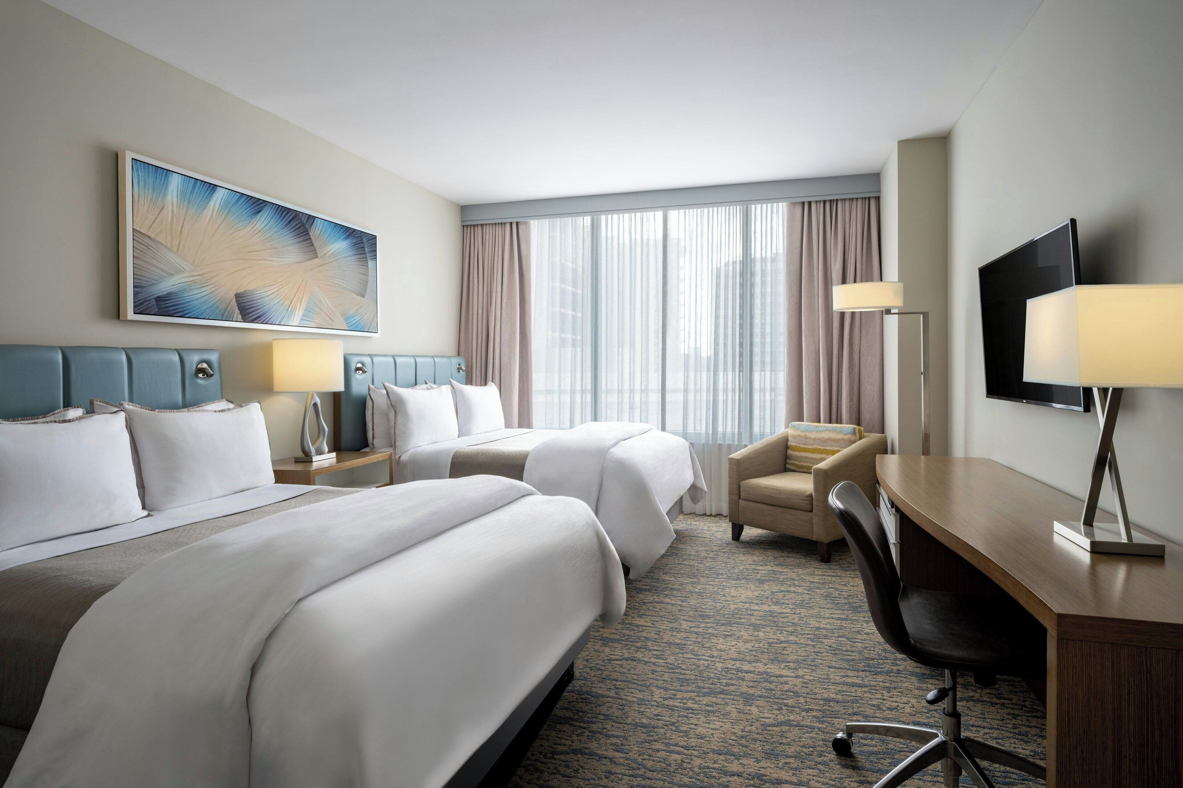 A hotel room with two beds, a desk, a chair, a TV, and a large window with curtains.