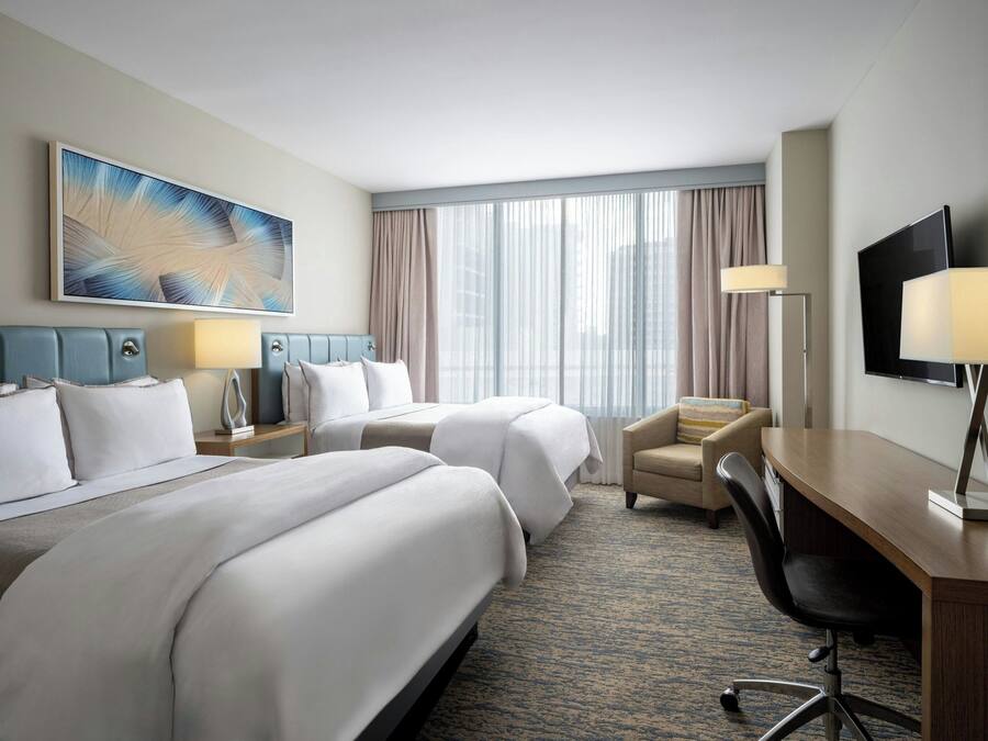 A hotel room with two beds, a desk, a chair, a TV, and a large window with curtains.