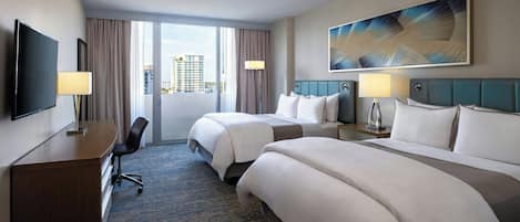 A hotel room with two beds, a desk, a TV, and a large window with a view of buildings.