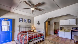 Un dormitorio con cama, ventilador de techo, puerta azul y una zona de cocina con armarios y electrodomésticos.
